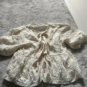 Krämvit blus med knyt från Gina Tricot - Supersöt krämvit blus från Gina Tricot i storlek XS. Blusen har en unik skrynklig struktur, voluminösa ärmar och snygga knytband framtill. Perfekt för dig som gillar romantiska och trendiga detaljer. Lätt och luftig känsla!