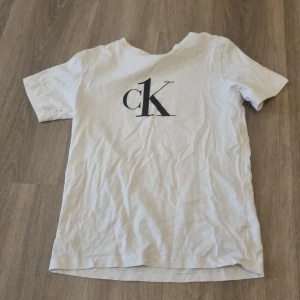 Vit Calvin Klein t-shirt med logga - Vit t-shirt från Calvin Klein med klassisk svart CK-logga på bröstet. Enkel och stilren design med rund hals och korta ärmar. Perfekt för dig som gillar minimalistisk och trendig streetstyle.