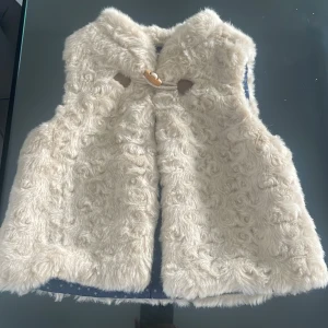 Krämvit pälsväst från Zara BabyGirl - Supermjuk och fluffig krämvit pälsväst från Zara BabyGirl. Västen har lockig fuskpäls, ett snyggt träknappsspänne och ett blått innerfoder med vita stjärnor. Perfekt för att ge outfiten en gullig och trendig touch.  storlek 86.  12-18 månader 