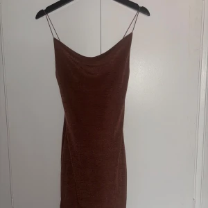 Brun kort klänning med tunna axelband - Säljer en snygg brun kortklänning med tunna axelband och lätt glansigt tyg. Klänningen har en enkel, rak passform och är ärmlös med en stilren look. Perfekt för dig som gillar minimalistisk stil och vill sticka ut med en varm brun färg.