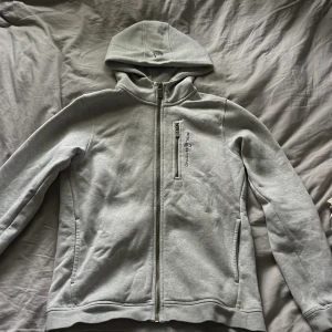 Ljusgrå hoodie från Sail Racing - Ljusgrå hoodie från Sail Racing med hel dragkedja, huva och bröstficka med dragkedja. Snygg logga på bröstet och huvan. Perfekt för chill dagar eller när du vill ha en clean look. Mjuk och skön bomullsblandning med långa ärmar. Hittade intw bild på den gråa