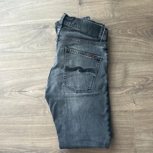 Grå jeans från Nudie Jeans Co - Snygga grå jeans från Nudie Jeans Co med klassisk femficksdesign och svarta broderade detaljer på bakfickorna. Jeansen har en rak passform och knäppning med knappar framtill. Perfekta för dig som gillar en stilren och avslappnad look. Använda men bra skick. Nypris 1800