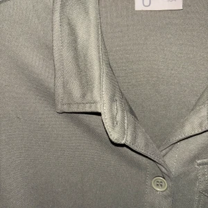 Olivgrön jumpsuit med knappar - Olivgrön jumpsuit från Uniqlo med skjortkrage, knappar framtill och knyt i midjan. Den har trekvartsärmar med knappdetalj och en bröstficka. Tillverkad i mjuk polyester som ger en avslappnad känsla.