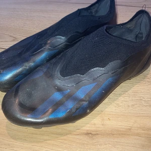 Adidas Crazyfast svarta fotbollsskor - Säljer ett par Adidas Crazyfast fotbollsskor i svart med blå detaljer och klassiska tre ränder på sidan. Skorna har en strumpliknande ovandel i syntetmaterial och mönstrad sula med fasta dobbar för bra grepp på planen. Snygg, slimmad design utan snörning.
