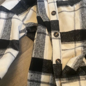 Rutig overshirt från SHEIN i svart och vitt - Snygg rutig overshirt från SHEIN i svart, vitt och grått. Skjortan har klassiska knappar framtill och är tillverkad i mjukt syntetmaterial. Perfekt att slänga över en t-shirt för en chill look.