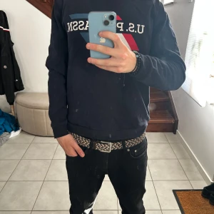 Blå sweatshirt U.S. Polo Assn. - Svart sweatshirt från U.S. Polo Assn. med tryckt logga och grafiskt mönster i rött, vitt och blått på bröstet. Tröjan har rund hals, långa ärmar och en normal passform. Perfekt för dig som gillar en clean och sportig stil.