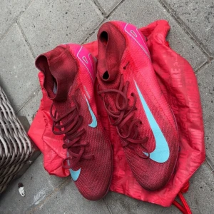 Nike mercurial elite  - Snygga fotbollsskor från Nike i rött med ljusblå swoosh och rosa detaljer. Ovandelen är i syntetmaterial med snörning och plös i samma färg. Sulan är rosa med blå inslag och har fasta dobbar för bra grepp på planen. Perfekta för dig som vill sticka ut på matchen!