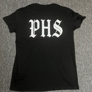 Svart PHS Support t-shirt - Svart t-shirt med stort PHS-tryck i vitt på ryggen och mindre PHS Support-logga framtill. Klassisk rund hals och korta ärmar. Skön passform och tillverkad i mjukt bomullstyg. Perfekt för dig som gillar streetwear och tydliga prints.