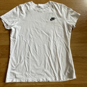 Vit Nike t-shirt med logga - Klassisk vit t-shirt från Nike med svart logga på bröstet. Enkel och stilren design med rund hals och korta ärmar. Perfekt till jeans eller shorts för en avslappnad look.
