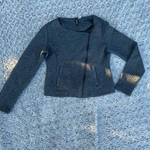 Grå quiltad jacka från Divided - Snygg grå jacka från Divided med quiltat sicksackmönster och asymmetrisk dragkedja. Jackan är figursydd, har rund halsringning och långa ärmar. Perfekt för dig som gillar en stilren look med lite edge.