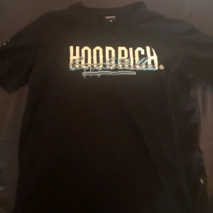 Svart t-shirt från Hoodrich M - Svart t-shirt från Hoodrich i storlek M med tryck i vitt och blått framtill och på ryggen. Snygg streetstil med coola texter och grafiska detaljer. Klassisk passform och rund hals. Perfekt för dig som gillar urban vibe och statement-plagg.