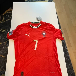 Portugal Ronaldo #7 fotbollströja Puma - Riktigt snygg Portugal fotbollströja från Puma med Ronaldo och nummer 7 på ryggen. Tröjan är röd med vita detaljer, har korta ärmar och broderat landslagsemblem. Kragen och ärmsluten har gröna och svarta ränder. Tillverkad i lätt och ventilerande material.