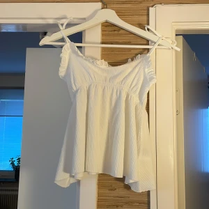 Vit volangtop från 157, stl S - Supersöt vit topp från 157 med volangkant och smala axelband som knyts. Luftig modell med rynkade detaljer och lätt struktur i tyget. Perfekt för varma dagar och ger en romantisk vibe till din outfit.