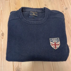 Mörkblå stickad tröja från Morris - Mörkblå stickad tröja från Morris med rund halsringning och ribbade muddar. På bröstet sitter ett snyggt tygmärke med Union Jack och texten 'Morris Gentlemen's Club'. Perfekt för dig som gillar klassisk och stilren look.