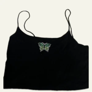 Svart linne med broderad fjäril - Svart linne med tunna axelband och broderad fjäril framtill. Fjärilen finns i olika färger, bland annat grön och rosa. Toppen har en enkel och stilren design som passar perfekt till jeans eller kjol. Materialet känns mjukt och bekvämt mot huden.