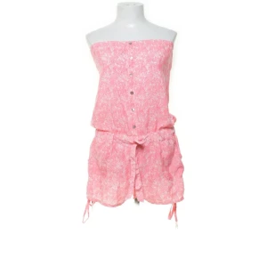 Rosa mönstrade byxdress från River Island - Snygga rosa byxdress från River Island med vitt blommigt mönster. Modellen är med knytband i midjan och knappar framtill. Lätta och luftiga, perfekta för varma dagar. Materialet känns mjukt och svalt mot huden.