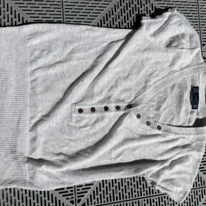 Ljusgrå stickad kortärmad tröja Zara - Superfin ljusgrå stickad tröja från Zara med korta ärmar och djup v-ringning. Tröjan har snygga knappar framtill och bred ribbad mudd nedtill. Perfekt till jeans eller kjol för en chill och stilren look.