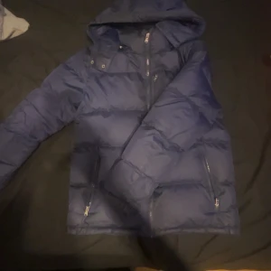 Blå dunjacka från Polo Ralph Lauren XL - Snygg blå dunjacka från Polo Ralph Lauren.Jackan har huva, dragkedjor på fickorna och quiltad design. Perfekt för kalla dagar med sin fluffiga och varma känsla. Klassisk look med diskret logga på insidan.
