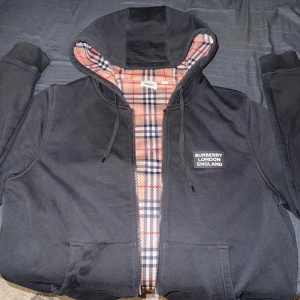 Svart Burberry hoodiejacka med rutig lining - Svart hoodiejacka från Burberry med dragkedja och klassiskt rutig lining i beige, rött och svart. Jackan har huva med snörning, två fickor fram och Burberry London England-tryck på bröstet. Perfekt för dig som vill ha en stilren streetlook med lyxig touch.