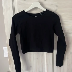 Svart långärmad croppad topp H&M Sport - Svart långärmad croppad topp från H&M Sport. Toppen har en enkel, stilren design med rund halsringning och lätt ribbad struktur. Perfekt för dig som gillar sportiga och minimalistiska plagg. Snygg att matcha med höga jeans eller tights.