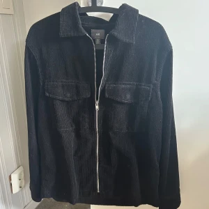 Svart manchesterjacka från H&M - Svart manchesterjacka från H&M i regular fit. Jackan har dragkedja framtill, två stora bröstfickor med lock och klassisk krage. Perfekt för dig som gillar en stilren och enkel look med lite edge.