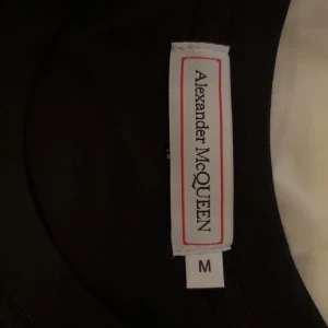 Svart t-shirt Alexander McQueen M - Svart t-shirt från Alexander McQueen i bomull med färgglad tryckt logga och texten 'EST. 1992' på bröstet. Klassisk rund hals och korta ärmar. Perfekt statement-plagg för dig som gillar designer och streetstyle. Storlek M helt ny