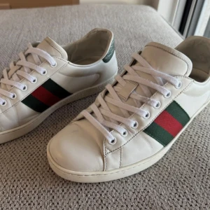 Gucci vita sneakers med röd-gröna ränder - Snygga vita sneakers från Gucci med klassiska röd-gröna ränder på sidorna. Skorna har rund tå, vit snörning och är tillverkade i skinn. Perfekta för dig som vill ha en stilren och lyxig look.