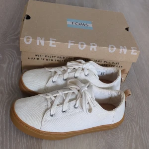 Vita sneakers från TOMS - Snygga vita sneakers från TOMS i canvas med klassisk snörning och ljusbrun gummisula. Skorna har rund tå och diskret TOMS-logga baktill. Perfekta för dig som gillar en clean och stilren look.