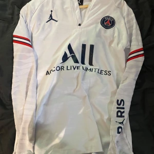 Vit långärmad PSG fotbollströja Jordan - Vit långärmad PSG fotbollströja från Jordan med klubbmärke på bröstet, mörkblå loggor och text, samt röda och blå ränder på ärmarna. Tillverkad i lätt och ventilerande material, perfekt för träning eller match. Snygg detalj med 'PARIS' på ärmen och 'VISIT RWANDA' på ryggen.