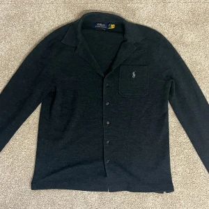 Mörkgrå merinoullsskjorta/cardigan Polo Ralph Lauren - Snygg mörkgrå skjorta/cardigan från Polo Ralph Lauren i mjuk merinoull. Klassisk krage, knäppning framtill och bröstficka med broderad logga. Långärmad och normal passform, perfekt för dig som gillar stilren och enkel design.