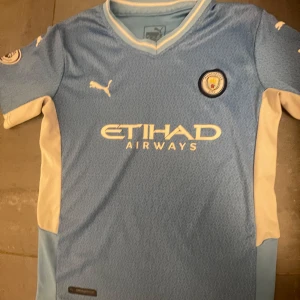 Manchester City hemmatröja Puma - Manchester Citys officiella ljusblå hemmatröja från Puma med klubbmärke på bröstet och Etihad Airways-tryck. Tröjan har korta ärmar, vita detaljer på sidorna och subtilt geometriskt mönster i tyget. Perfekt för dig som älskar fotboll och vill visa ditt stöd för City.