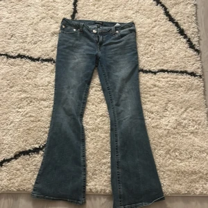 True Religion bootcut jeans blå, stl 29 - Säljer ett par klassiska blå jeans från True Religion med bootcut passform. Jeansen har snygga kontrastsömmar och ikonisk broderad bakficka. Tillverkade i jeansmaterial med en cool tvättad look. Perfekta för dig som gillar en avslappnad men trendig stil. Dock klippta inuti för att dom var för stora (ditsatta med band) inget som syns utanför byxan