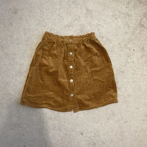 Brun manchesterkjol från H&M - Säljer en brun kort manchesterkjol från H&M i storlek 122/128. Kjolen har knappar framtill, resår i midjan och är i ett mjukt, ribbat tyg. 