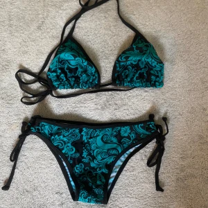 Svart och turkos bikini med knyt - Snygg bikini med svart bas och turkost blommigt mönster. Triangeltopp med knyt i nacken och ryggen, samt matchande bikinitrosa med knyt i sidorna. Perfekt för stranden eller poolen. Materialet känns som syntet och torkar snabbt.