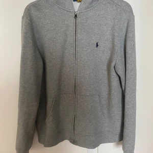 Grå hoodie från Ralph Lauren - Klassisk grå hoodie från Ralph Lauren med dragkedja framtill och broderad logga på bröstet. Tröjan har huva, långa ärmar och två fickor framtill. Perfekt för en chill och stilren look.