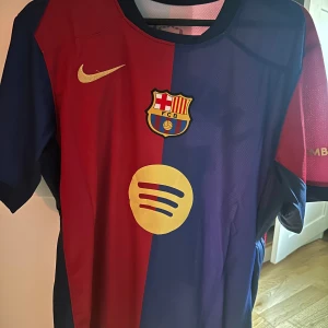 FC Barcelona Lamine Yamal matchtröja - Säljer en officiell FC Barcelona matchtröja från Nike med Lamine Yamal och nummer 19 på ryggen. Tröjan har klassisk röd och blå design, korta ärmar och klubbmärke på bröstet. Spotify-logga på framsidan och UNHCR-detalj på ryggen. Perfekt för dig som älskar Barca!