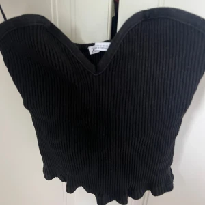 Svart ribbad tubtopp från Zara - Svart ribbad tubtopp från Zara med söt vågig nederkant och hjärtformad urringning. Toppen är ärmlös och har en croppad passform som sitter snyggt på kroppen. Perfekt att styla med jeans eller kjol för en trendig look.