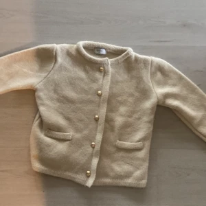 Beige stickad kofta från Rihoas - Superfin beige stickad kofta från Rihoas med guldknappar framtill och två små fickor. Klassisk rund halsringning och långärmad modell. Perfekt för lager-på-lager och enkel att styla till olika outfits.