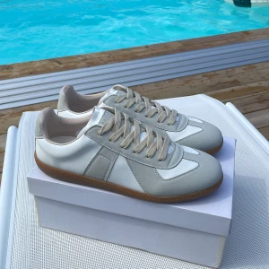 Maison Margiela sneakers  - Maison Margiela sneakers i vitt skin och ljusgrå mocka. Skorna är helt oanvända. Skorna är små i storleken så dessa skor passar 42. Box samt dustbags medföljer. Hör av er vid frågor eller intresse! Skickar samma dag!(Obs inte 0rginal)