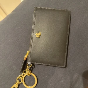 Svart RL plånbok i skinn med gulddetaljer - Snygg svart plånbok från Ralph Lauren i slätt skinn med guldfärgat RL-logo framtill. Plånboken har dragkedja, flera kortfack på baksidan och guldfärgade detaljer som nyckelring och karbinhake. Perfekt för kort, mynt och nycklar. Helt ny aldrig använd! Köptes för 1200kr säljer för 800
