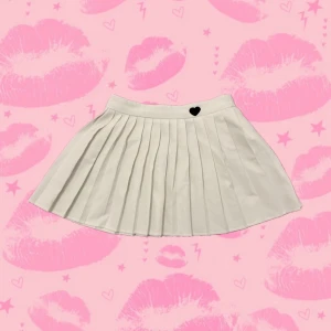 Vit plisserad kjol från SHEIN XS - Supersöt vit plisserad kjol från SHEIN i storlek XS. Kjolen har klassiska veck och en liten svart hjärt-detalj i midjan. Tillverkad i bomullsblandning för en skön känsla och snygg passform. Perfekt för dig som gillar preppy och trendig stil. 