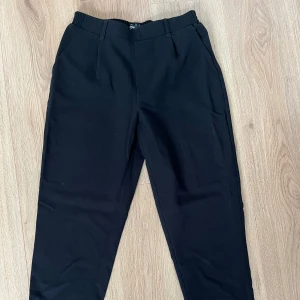 Svarta kostymbyxor med resårmidja - Svarta kostymbyxor med elastisk midja och hällor för bälte. Klassisk rak passform med diskreta fickor på sidorna. Perfekta för en clean och stilren look. Materialet känns mjukt och följsamt, troligtvis polyesterblandning.