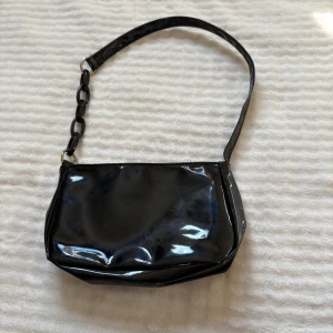 Svart glansig axelväska med kedjedetalj - Snygg svart axelväska i glansigt syntetmaterial med cool kedjedetalj på axelremmen. Väskan har en enkel, stilren design och passar perfekt när du vill addera lite edge till din outfit. Rymmer mobil, plånbok och andra småsaker.