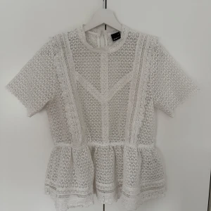Vit spetsblus från Gina Tricot - Superfin vit blus i spets från Gina Tricot med korta ärmar och rund hals. Blusen har ett vackert mönster med detaljerade broderier och volang nedtill. Perfekt för dig som gillar romantisk och feminin stil.