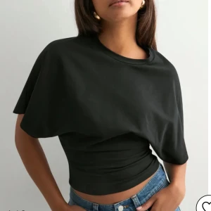 Svart croppad topp från Nelly - Svart croppad trendig topp från Nelly med unik lager-på-lager look. Toppen har korta, vida ärmar och sitter tight runt midjan för en snygg siluett. Perfekt att matcha med jeans eller kjol för en cool och trendig stil.