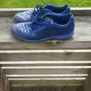 Blå Nike fotbollsskor med snörning - Snygga blå fotbollsskor från Nike med quiltad ovandel och svarta detaljer på sidorna. Skorna har snörning och låg profil, perfekt för dig som gillar att spela inomhus eller på konstgräs. Röd innersula och klassisk Nike-logga på sidan.