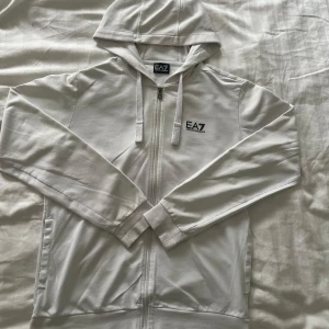 Vit ziphoodie från EA7 Emporio Armani - Snygg vit hoodie med dragkedja från EA7 Emporio Armani. Jackan har huva med snörning, svart logga på bröstet och diskreta ränder baktill. Tillverkad i mjuk bomullsmix, perfekt för en clean och sportig look.