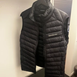 Svart dunväst Peak Performance - Säljer en svart dunväst från Peak Performance med quiltad design och hög krage. Västen har dragkedja framtill och logga broderad på bröstet. Perfekt att slänga över en hoodie för extra värme. Klassisk och stilren look.