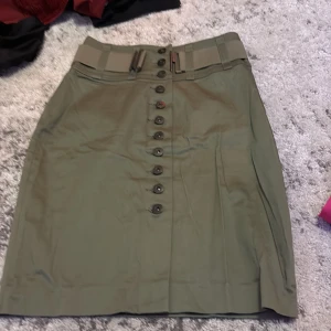 Olivgrön kjol med knappar Gina Tricot - Snygg olivgrön kjol från Gina Tricot med hög midja och bred linning. Kjolen har dekorativa knappar framtill och ett brett bälte med spänne. Rak passform och stilren look, perfekt till sneakers eller boots.