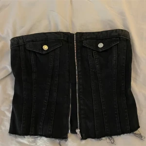 Svart jeanstopp från Zara - Cool svart korsettopp från Zara i jeansmaterial med dragkedja framtill och råa kanter nertill. Toppen har två dekorativa fickor med silverknappar och markerade sömmar som ger en edgy vibe. Perfekt för att lyfta vilken outfit som helst.